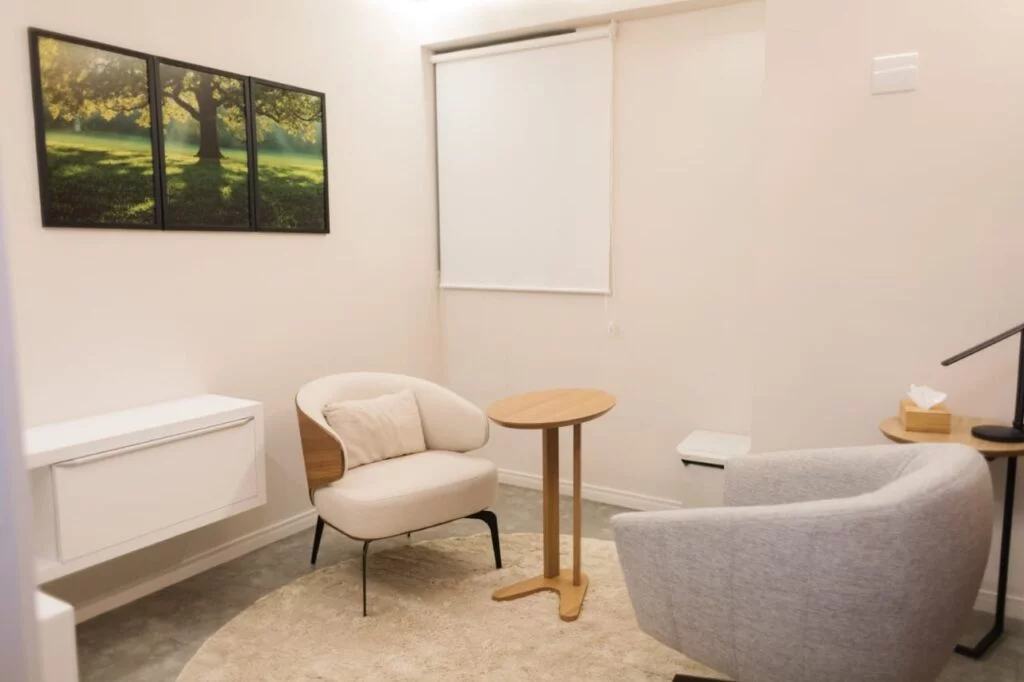 Uma sala de psicologia para atendimento psicológico com duas poltronas beges, uma mesa de centro e armário, uma janela com cortinas brancas e um quadro na parede.