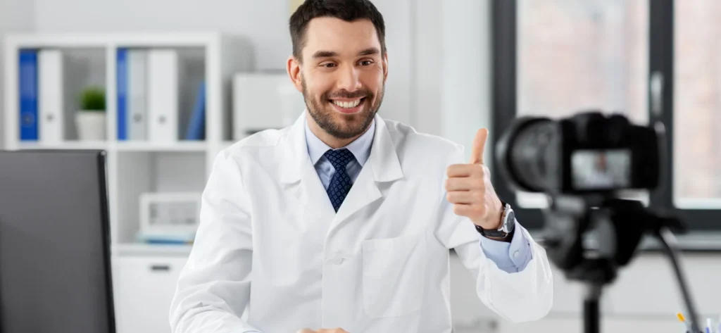 Um médico sorridente vestindo um jaleco branco e gravata está dando um sinal de "joinha" enquanto olha para a câmera de vídeo em um consultório médico claro e moderno.