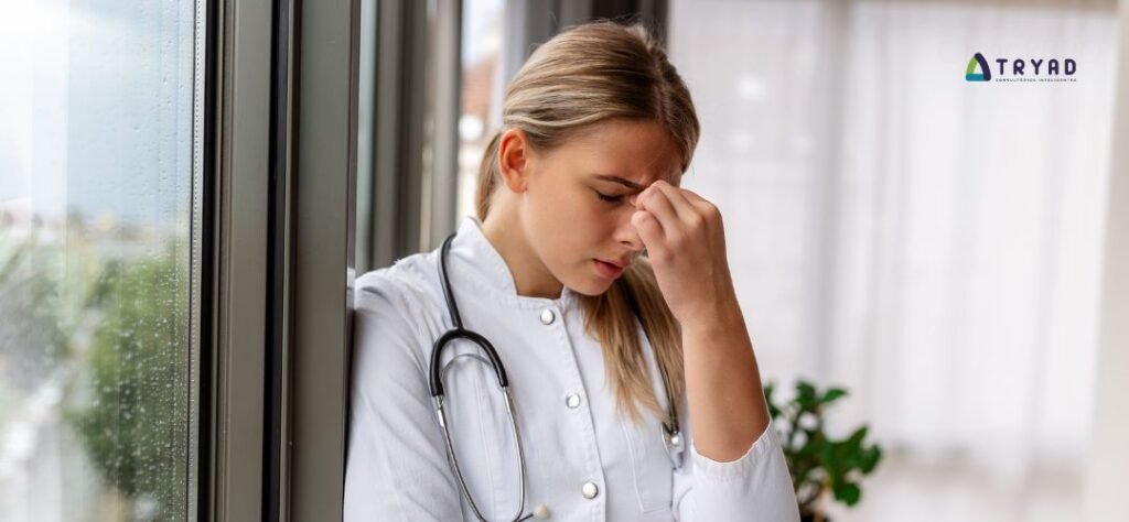 Médica de jaleco branco com expressão de cansaço ao lado da janela em ambiente clínico.