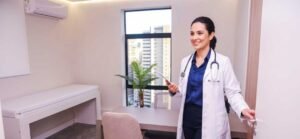 Médica sorridente entrando em consultório equipado com maca, cadeiras e vista para a cidade, representando o atendimento em consultório compartilhado em Recife